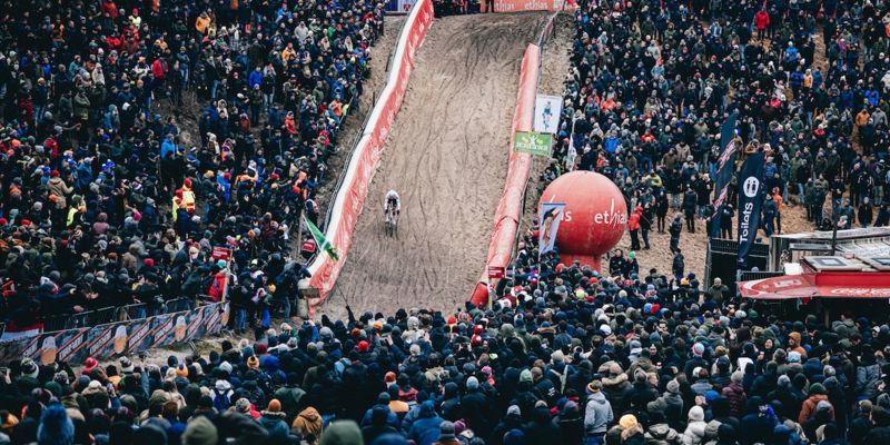 Crowds of spectators watch cyclists racing up a steep muddy hill during a cyclo-cross event.