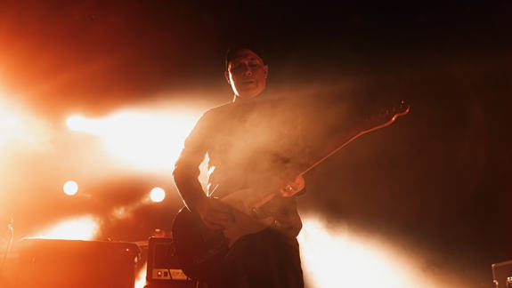 Stuart Braithwaite A musician silhouetted by bright orange stage lights plays an electric guitar amidst smoke or haze.