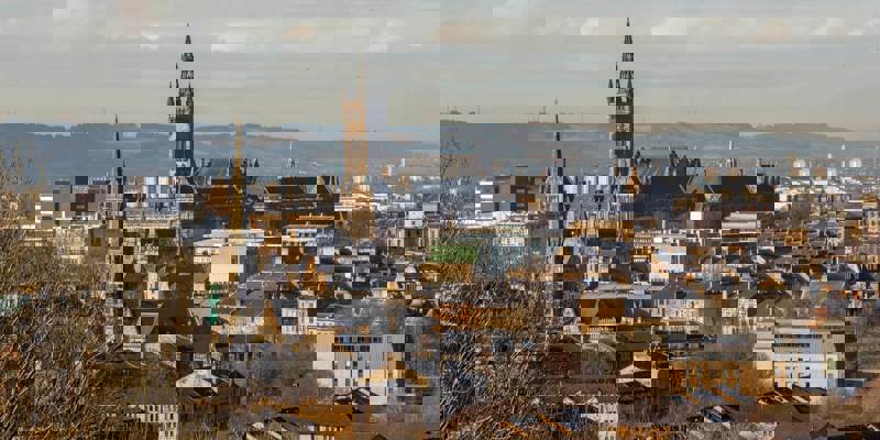 An aerial view of Glasgow's cityscape with the University of Glasgow building prominent in the centre, seen from a hillside. The foreground is blurred with brown and green brush, while a hilly horizon is visible in the distance.