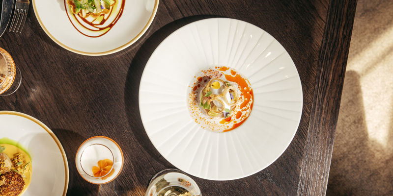 Overhead shot of small, elegantly plated dishes on a dark wooden restaurant table, with wine glasses.