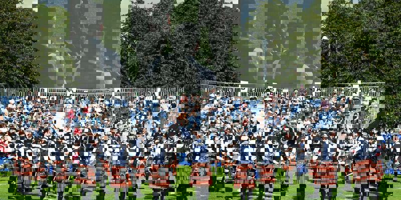 A group of people playing bagpipes and drums in a circle on a grassy field in front of a crowd and a large video screen.