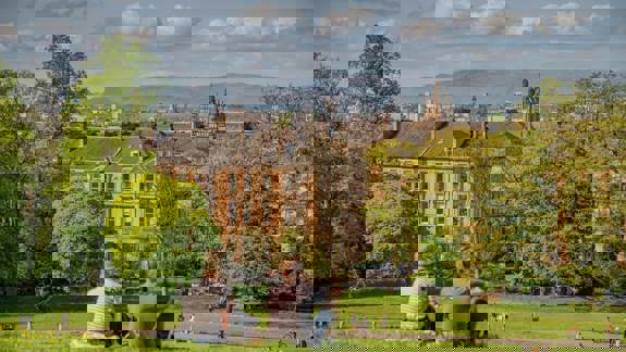 Two people sit on a grassy hill, looking out at a city skyline with buildings and distant hills under a cloudy sky.