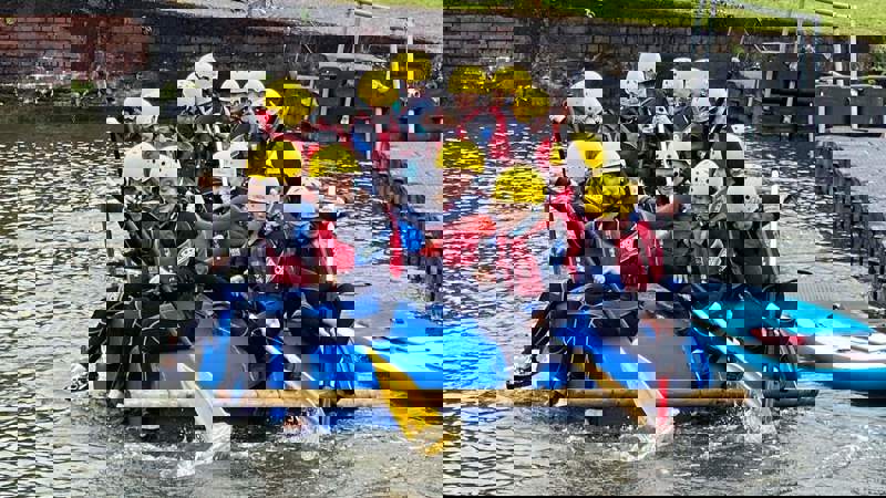 A group of children in wetsuits and yellow helmets paddling a raft made of blue barrels on a body of water.