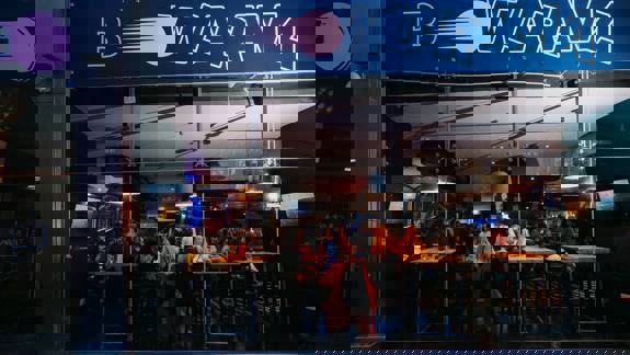 Exterior view of Bowlarama bar and bowling venue at night, showing people sitting at tables inside through large glass windows.