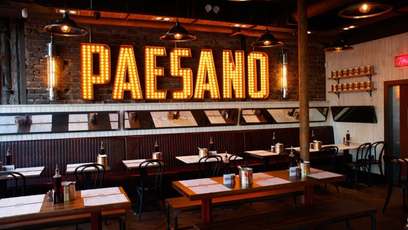 Interior of a restaurant with rustic seating, long tables, and a large "PAESANO" sign with exposed light bulbs.
