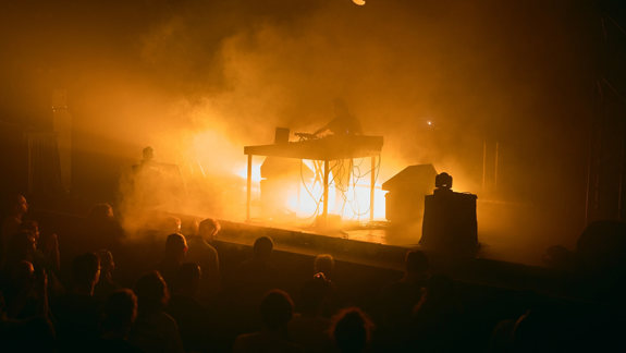 An atmospheric photo of a musician performing an electronic set on a dimly lit stage, engulfed in thick fog and intensely bright, warm orange light.
