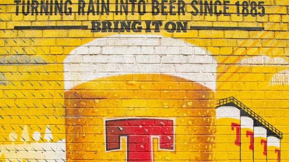 A vibrant yellow brick wall mural depicting a large beer glass with "Tennent's" and "Turning Rain into Beer Since 1885".