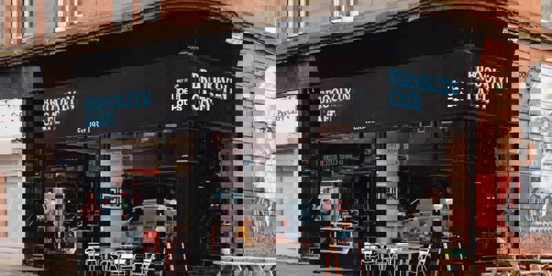 The black-fronted Brooklyn Cafe on a street corner with large windows and outdoor tables. A mural of a man can be seen on the corner of a sandstone building.