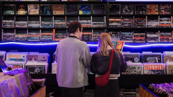 Two people standing with their backs to the camera, looking through vinyl records displayed in bins and on shelves in a record store lit with blue neon light.