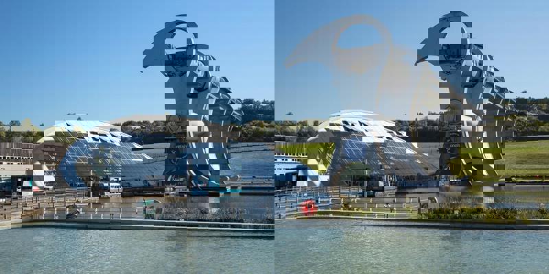 The Falkirk Wheel, a huge, modern rotating boat lift, sits next to a glass-and-steel building on a sunny day.