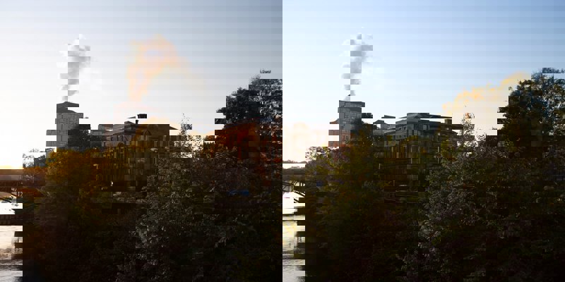 A large, historic red brick building by a river at sunrise, framed by dark trees. Smoke rises from a distant tower.