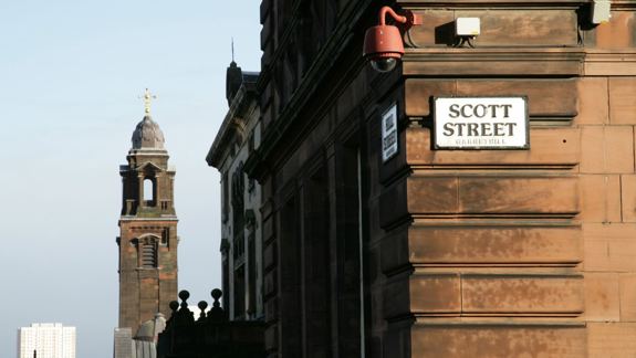Street scene with a building on the right labelled "Scott Street." A historical tower with a cross rises in the background. Bright, clear sky above.