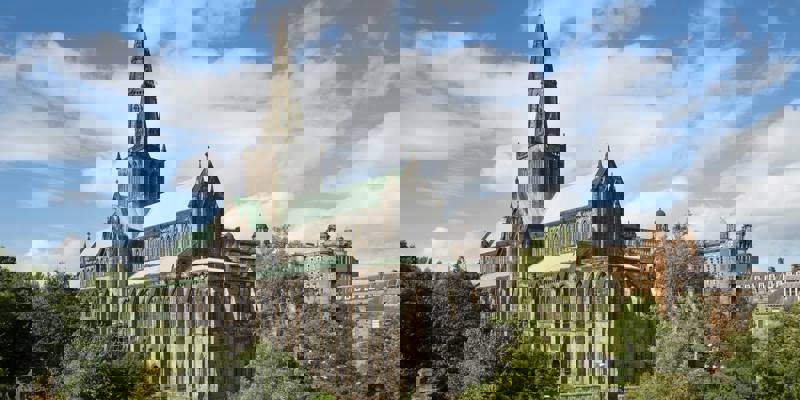 The grand exterior of Glasgow Cathedral with its spire reaching into a blue sky with white clouds, surrounded by green trees.
