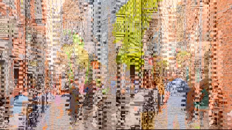 A bustling cobbled lane with outdoor dining, string lights, and historic brick buildings under a bright sky.