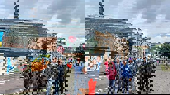 A large group of people smiling on a sidewalk in front of a brick building with a large mural and a steeple.
