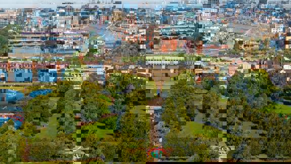 An aerial view of Glasgow Green, showing expansive green spaces, tree-lined paths, a colorful sculpture, and the River Clyde with bridges in the background.