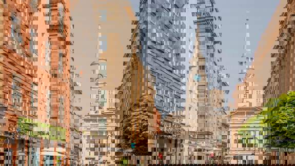 A wide street view showing historic architecture in Glasgow, including a church with a tall steeple and clock face, and several cafes.