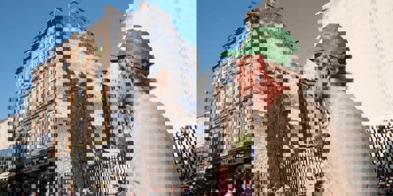 A person in a green baseball cap looks towards a large mural covering the gable end of a building. The mural is of a person in a winter coat, one arm raised high towards the sky