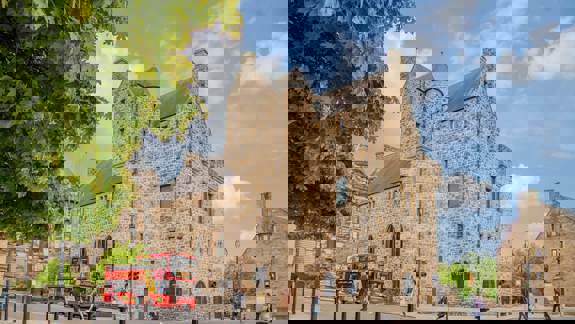 A large, stone building with a castle-like appearance stands on a street corner with a red double-decker bus passing by.