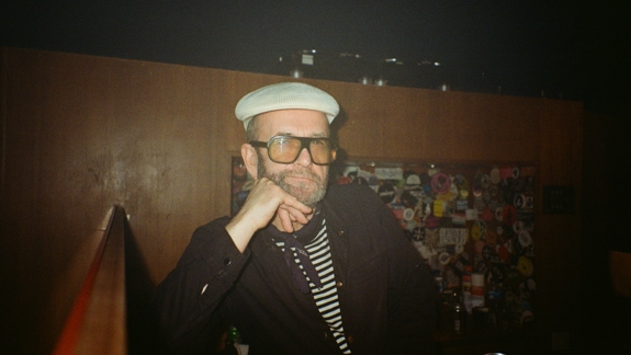 A man in a flat cap and striped shirt, wearing large glasses, sits at a bar with his hand on his chin.