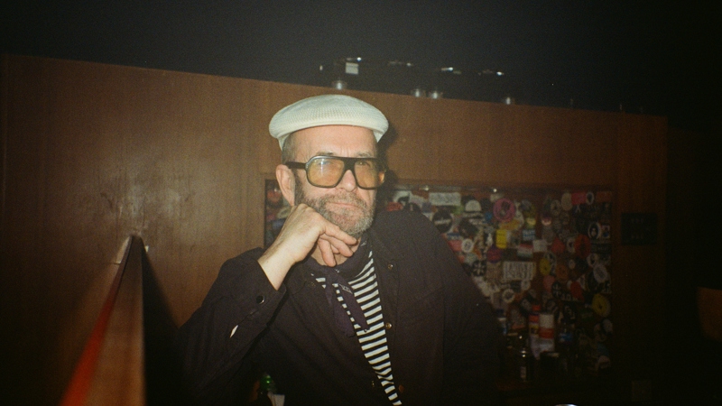 A man in a flat cap and striped shirt, wearing large glasses, sits at a bar with his hand on his chin.