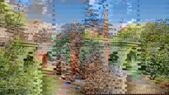Historic stone and iron bridge over the River Kelvin in Glasgow, surrounded by lush greenery, with cyclists on the riverside path and a church spire in the background.