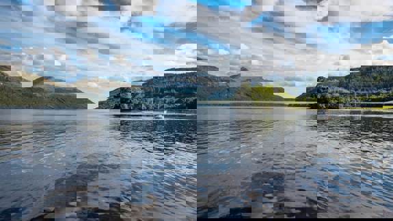 A large body of water surrounded by mountains and hills under a cloudy blue sky.