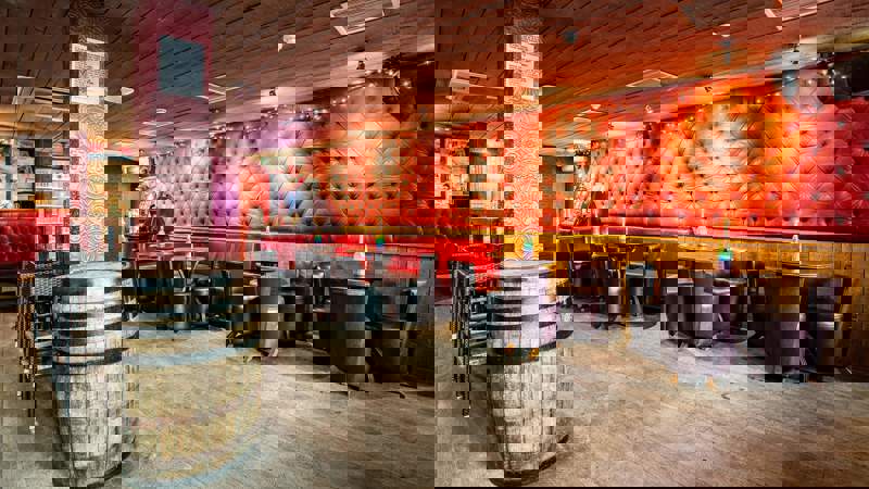 An empty speakeasy-style bar with red-tufted leather banquettes, wooden floors, and a prominent wooden barrel.