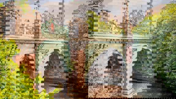 A picturesque stone and iron bridge over a river, with two people walking on the riverside path and a church spire rising behind trees in the distance.