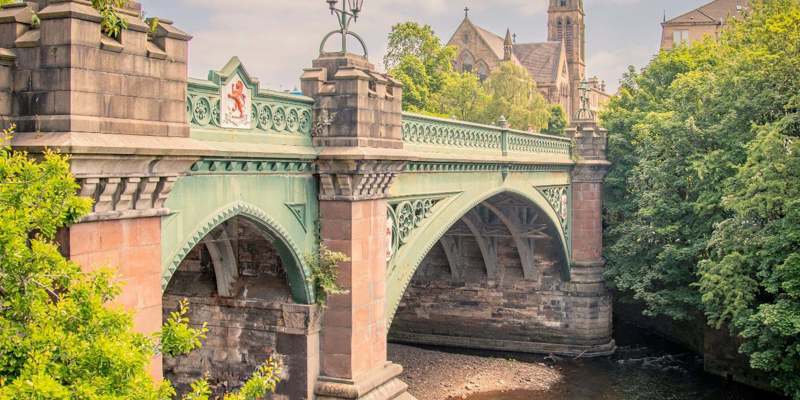 A picturesque stone and iron bridge over a river, with two people walking on the riverside path and a church spire rising behind trees in the distance.