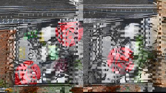 Colourful mural on a brick wall featuring bold geometric patterns and red roses, inspired by stained glass design, created by street artist Rogue-One.