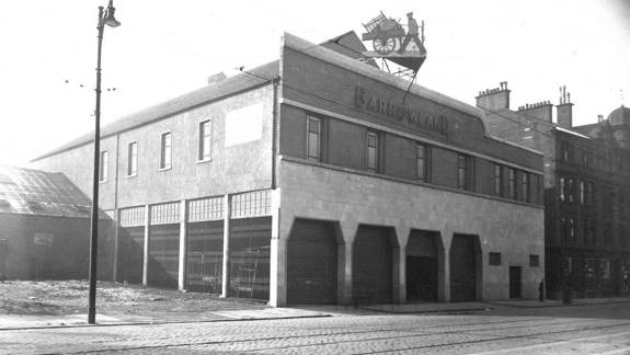 A black and white historical photo of the original Barrowland building, showing a brick and stone facade and an antique cart statue mounted on the roof.