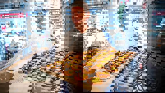 A baker in a white uniform smiles while proudly holding a large tray of freshly baked, traditional Portuguese Pastéis de Nata in a bakery.