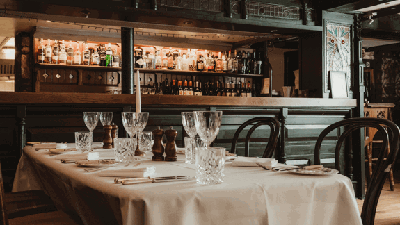 Elegant restaurant table with white tablecloth, crystal glasses, cutlery, napkins, salt and pepper shakers, and background bar with wooden shelves and bottles.