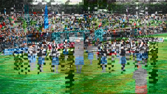 A wide shot of a pipe band competition with many bandsmen in kilts playing bagpipes and drums in a grassy field.