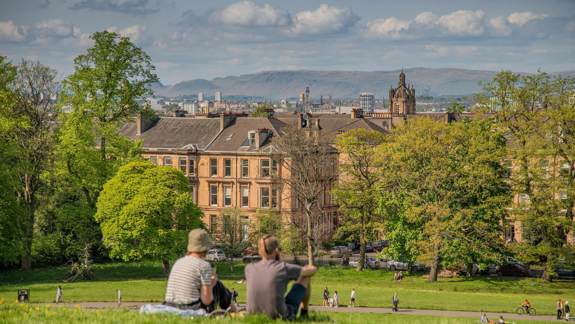 Two people sit on a grassy hill, looking out over a park and a city skyline with hills in the distance.