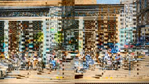 People are seated at outdoor tables in front of a café with large windows on a sunny city street.