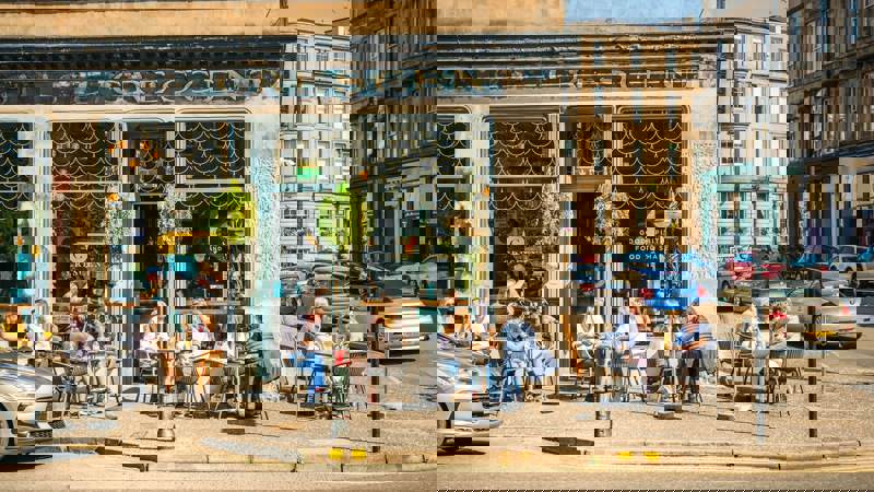 People are seated at outdoor tables in front of a café with large windows on a sunny city street.