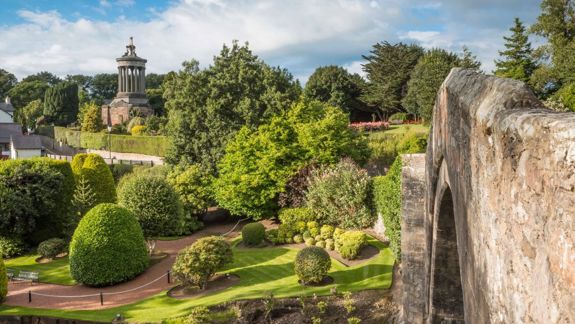 A scenic view of the Burns Monument in Alloway, Scotland. The monument, a classical temple on a hill, is surrounded by lush gardens and trees, with an old stone bridge in the foreground.