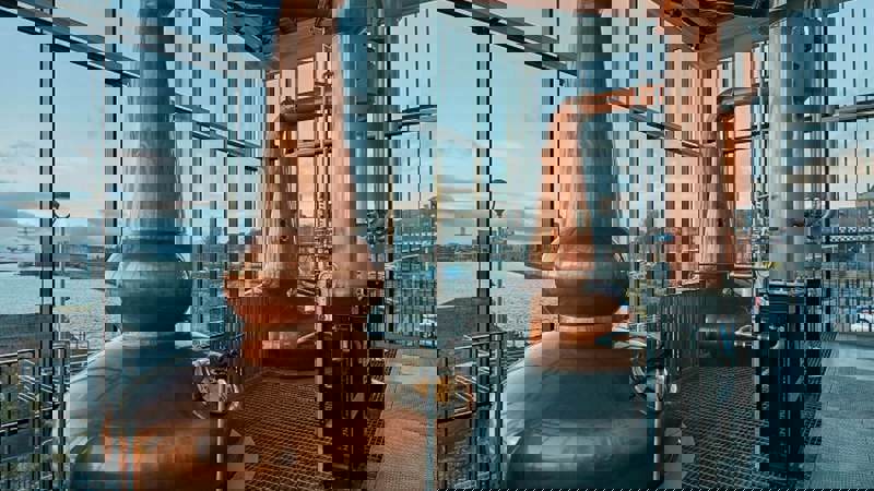 A modern distillery with two large, shiny copper stills and large windows looking out onto water.