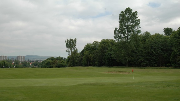 A wide green golf course with a red flag on the putting green, bordered by tall trees and city buildings visible in the distance.