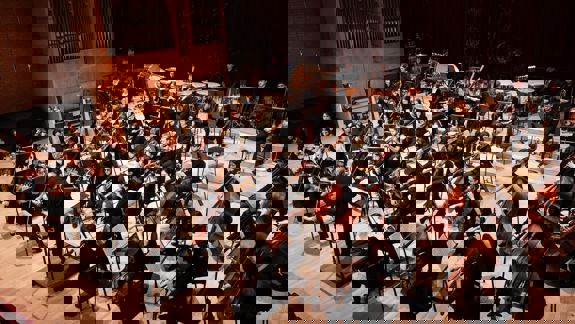 A large orchestra performs on a stage, with a conductor facing the musicians and a large pipe organ in the background.