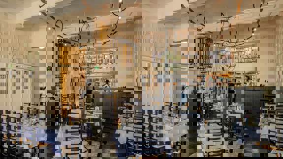 Ground-floor view of a bright, open restaurant interior featuring dark wooden chairs, white tablecloths, and decorative lighting fixtures. A staircase leads to an upper dining level in the background.