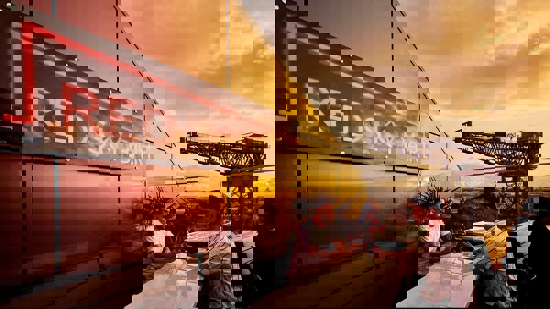 Two men are seated at a white table on a rooftop bar. The red neon sign for "RED SKY BAR" is prominent. A large crane is visible in the background.