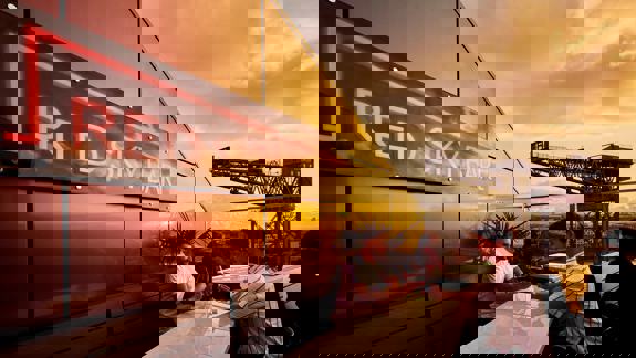 Two men are seated at a white table on a rooftop bar. The red neon sign for "RED SKY BAR" is prominent. A large crane is visible in the background.