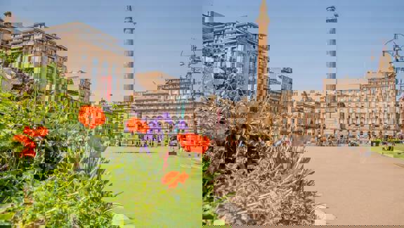 A sunny, wide-angle shot of a city square with a monument, surrounded by large buildings and flowers in the foreground.