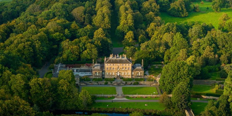 An aerial view of Pollok House within Pollok Country Park. The building is surrounded by lots of trees and bushes. The grounds are immaculately kept.