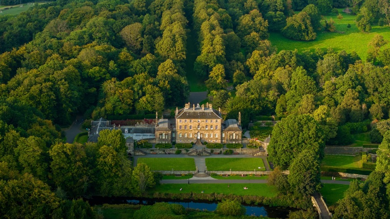An aerial view of Pollok House within Pollok Country Park. The building is surrounded by lots of trees and bushes. The grounds are immaculately kept.