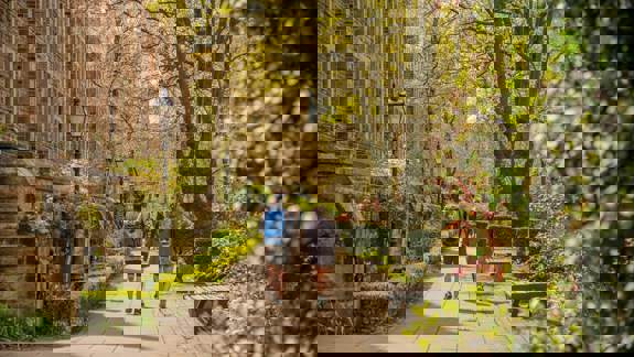 Two people walk down a paved pathway lined with benches, lush green trees, and a traditional stone building on a sunny day.