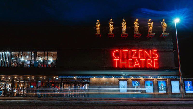 Exterior of Citizens Theatre in Glasgow at night with bright red neon sign, illuminated statues on the roof, and crowds visible through large windows on the left.